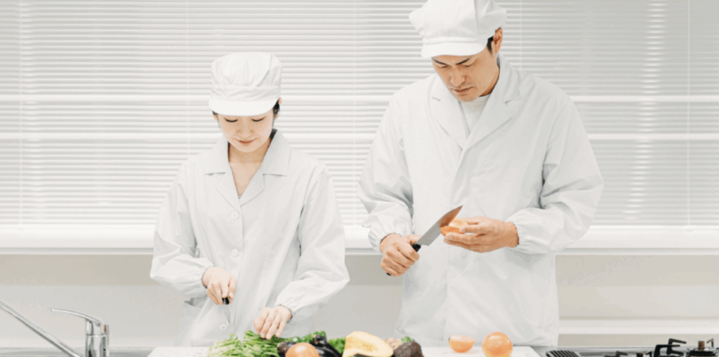 白衣と帽子を着用した男女の調理スタッフが厨房で野菜と玉ねぎを切っている