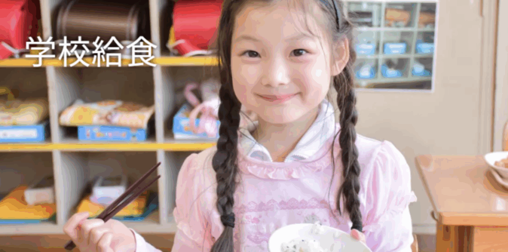 教室のロッカーを背景に、茶碗のご飯と箸を持ってほほ笑む三つ編みの小学生(左上に「学校給食」の文字)