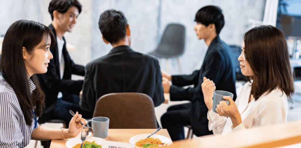 オフィスのカフェテリアで食事とコーヒーを手に会話を楽しむ複数の社員